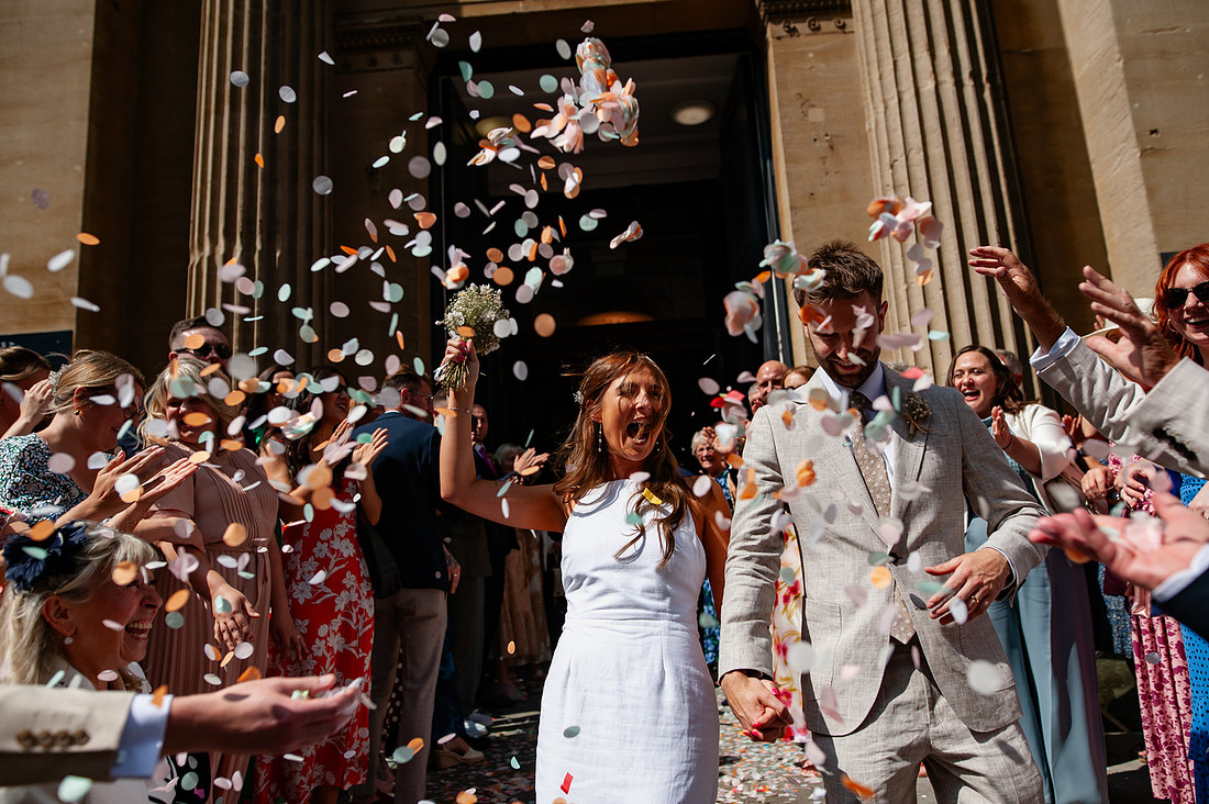 Bride and groom walking through confetti outside Bristol Registry Office while guests celebrate around them during a relaxed documentary-style wedding moment.