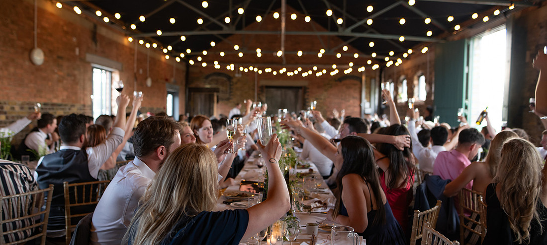 Wedding guests raising glasses during speeches inside a warmly lit wedding reception with fairy lights overhead.